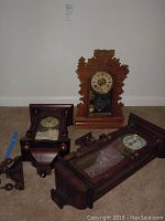Three vintage wooden wall clocks on floor: one carved, one with glass and columns, one simpler handmade style, all untested.