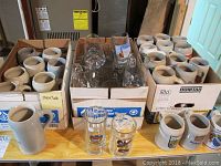 Overview photo showing multiple ceramic beer steins packed in 3 boxes along with two glass beer mugs positioned in front on the table.