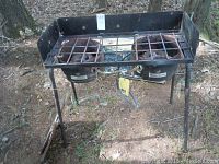 Outdoor propane stove top with two burners and metal grates showing rust and dirt, placed on natural ground.