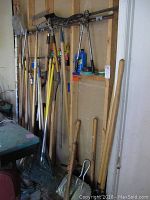 View of large assortment of long-handled garden tools including rakes, post hole digger, pick axe, and shovel leaning against wall.