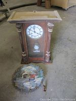Wood mantel clock with turned wooden columns, Roman numeral clock face, pendulum, and glass front standing upright on floor.