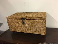 Closed wicker rectangular box with black metal latch, brown woven wicker material, placed on a wooden surface with white wall background.