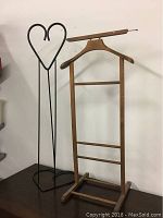 Photo of a wooden clothes rack narrow frame with hangers and a black metal heart-shaped stand set on a tabletop against a white wall.