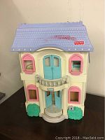 Closed view of Fisher Price doll house showing purple roof, cream walls, pink window shutters, teal front doors, green shrubbery on either side.