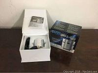 PowerPod 360 vibration speaker in silver color inside white open box with USB cable and instruction manual next to the retail box on a brown surface