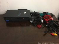 Photo showing Sony Playstation 2 console with controllers and cords on a wooden surface.