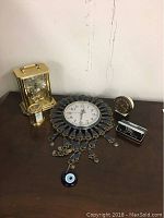 Six items on wooden surface including two gold-tone brass clocks, a decorative hanging clock with blue and gold accents, a small round clock, and Ekso binoculars in case