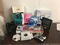 Photo showing variety of boxed electronics including Vomo speaker, Logitech webcam, digital cameras, Tivax TV antenna, flashlights and voice recorder