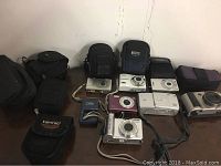 Seven cameras including film and digital on table with eight camera cases and Canon battery charger visible around