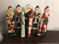 Photo showing nine wooden Santa Claus figures of varying height and designs, arranged on a wooden surface against a plain background.