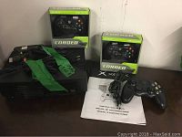 Original Xbox console with green label on top, two Pelican corded Xbox controllers in box, one loose controller, Xbox manual, and cables on table.