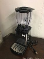 Full view of the smoothie maker showing the clear blending container with a lid and spout, and the black and stainless steel base with drip tray.