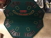 Top view of foldable octagonal poker table with green felt playing surface. The table has printed card positions for nine players, text 'CASINO DEALER'S CHOICE PLACE YOUR BETS' in red and gold, padded armrests with built-in plastic cup holders and chip trays around the perimeter.