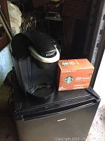 Black Keurig coffee maker with silver handle on top of black Galanz appliance, with box of Starbucks K-Cup pods beside it.