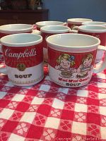 Photo showing 8 Campbell's Soup mugs in two sizes on red and white checkered cloth, clean, no chips visible