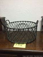 Full view of round wrought iron basket with lattice wire sides and metal handles sitting on wooden surface.