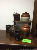 Six stoneware items displayed on a wooden table and a small shelf, showing a mix of brown and two-tone crock items with visible signs of wear and chips.