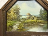 Close up of hand painted landscape showing cabin, river, trees, and sky in wooden frame.