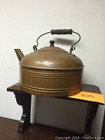 Side view of large copper kettle showing cylindrical body, black knob lid, long narrow spout, and wooden handle grips.
