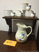 Four white ceramic soup bowls featuring goose head handles with blue bows, stacked on wooden shelf behind the pitcher.