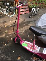 Photo showing front and side view of pink Razor electric scooter with padded seat, in outdoor setting with asphalt ground and garden in background.