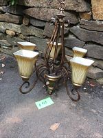 View of entire five-arm chandelier lamp including metal frame and five glass shades, placed in front of stone wall outdoors.