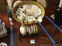 Photo showing large oval decorative plate with olive branch design and black metal stand, along with four matching saucers and ceramic bottle.