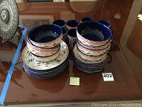 Photo of stacked plates and bowls with floral/leaf pattern and blue rim on table surface.