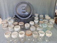 Wide view showing multiple Mason jars in clear and blue-tinted glass with metal lids arranged in front of a black speckled canning kettle with lid.
