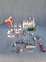 Overview of lot contents showing various bottle openers, bitters bottles with display box, corkscrews, silver-plate ashtray, whisky bottles, glass mug, and boxed shot glasses.