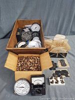 Wooden box filled with round meters and gauges, black analog multimeter, and numerous small wooden resistors in separate box; some loose signed gauge faces on table.