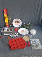 Photo showing various baking items including apple peelers, pie plates, muffin tins, foil, and baking molds arranged on a cloth background.