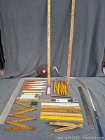 Full layout of collection showing yardsticks standing upright in back, multiple folding rulers arranged on a fabric surface, metal ruler with handle, and coiled tape measure.