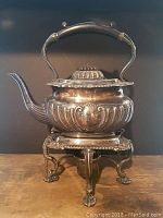 Front view of the silver plated tea pot resting on the ornate warming stand with claw feet.