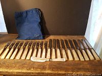 Full set of 10 forks, 10 knives and decorative butter knife with cloth bag behind them on wooden table.