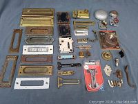 Photo showing a variety of door hardware components including letter slots, mortise locks, door bells, a door handle, and other metal pieces spread on a blue cloth background.