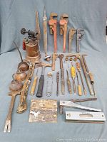 Full view of all tools arranged on a cloth backdrop showing a variety of vintage hand tools including brass blow torch, pipe wrenches, hand files, saw blade, scrapers, and staple gun.