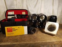 Variety of camera lenses, carrying case, Kodak lens cleaning paper and magnifying loupe arranged on wood surface