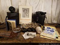 Wide shot of all items spread on table including the Print Finishing book, vintage camera lens and case, photo album, colorful woven strap, key holder, lens caps, flash bulb with cord, small photos, and brochures.
