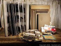 Photo showing large black and white photo album with thick pages and numerous loose vintage photographs along with photo mounting supplies