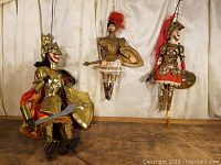 Three Italian soldier marionettes displayed against a curtain background, showing one larger and two smaller puppets in detailed costumes with swords and shields.