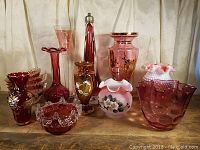 Front view displaying various cranberry glass pieces including tall lamp base, vases, bowls, and dishes in different shapes and decorated styles.