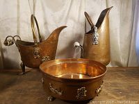 Four copper decorative items are shown including a coal scuttle, tall pitcher, footed oval planter, and a small round bowl all with brass and porcelain decorative elements. Pieces have a hammered texture and polished finish.
