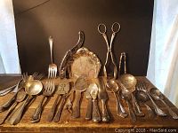 Overall view of an assortment of silver-plated service implements laid out on a wooden surface showing spoons, forks, knives, tongs, and a decorative tray in the background.