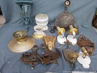 Overview of vintage lighting fixtures including two white ceramic bases, multiple metal wall sconces in brass and bronze, a large brass round lamp shade, and a patina copper outdoor lantern.