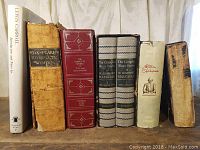 Seven books placed upright on a wooden surface against a cream curtain. From left to right: The Annotated Alice, vintage worn tan book titled Shakspeare's Complete Works, red leather bound Complete Works of William Shakespeare, two volumes of The Complete Short Stories of W. Somerset Maugham, a white book with Shakespeare illustration, heavily worn book on far right.