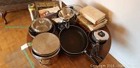Photo showing the entire lot on a round wooden table including pots with lids, a fry pan, a steamer insert, an electric waffle iron, and multiple books stacked beside the cookware.