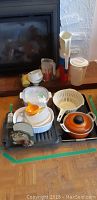 Photo of kitchen items including Corningware orange enamel pot, white enamelware pot, colander, measuring cups, jugs, and utensils arranged on floor.