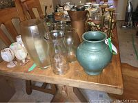 Photo showing assortment of glass and ceramic vases of various sizes on wooden table, including green ceramic vase, frosted white glass vases, clear glass vases, and brass candlestick holders.