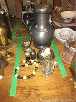 Photo showing a 10" Dutch pewter jug with stamp on bottom alongside two small brass and blue glass salt and pepper shakers, multiple candle holders, a bracelet, and a shot glass on a wooden table.
